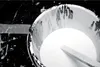 A close-up, high-angle shot of a white bucket filled with white liquid, with a wooden stick resting inside. The bucket is splattered with white paint, and the background is black with white splatters and a strip of grey tape. Anteprima dell’immagine prodotto