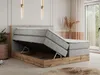 A modern bedroom with a king-size boxspring bed with storage, featuring a lifted mattress to reveal the storage compartment. The bed has a light grey upholstered headboard and frame, with a wooden base in Wotan oak finish. The room is decorated with minima Produktbild-Vorschau