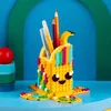 A LEGO DOTS banana-shaped pencil holder with pens and pencils inside, displayed on a blue surface against a blue and green background. Podgląd zdjęcia produktu
