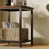 A close-up view of a square side table with a wooden top and black metal frame, featuring a woven basket filled with books on the lower shelf. Produktbild-Vorschau