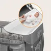 A baby is lying on a changing pad attached to a baby playpen. The playpen has a grey and white patterned fabric and storage compartments. Produktbild-Vorschau