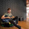 A man is sitting on a bean bag chair and playing a guitar in a room with a brick wall. Voir les images du produit