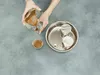 Hands pouring a brown substance from a glass container into a small glass, next to a stainless steel pot with a perforated insert and two covered containers. Produktbild-Vorschau
