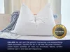 A close-up of a large white pillow on a bed, with a grey blanket draped over it. A circular gold and black badge with text is on the right side of the pillow. Produktbild-Vorschau