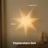 A large paper star lantern hangs in the center of the frame, illuminated from within. To the right, a small, folded paper Christmas tree decoration sits on a surface. The background is a plain, light-colored wall. Podgląd zdjęcia produktu