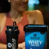 A person is in the background, blurred, behind a protein shaker and a tub of whey protein. Produktbild-Vorschau