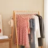 A wooden clothing rack with various garments displayed on pink velvet hangers, including a pink polka dot dress, a plaid top, and jeans. A beige knitted hat hangs on the side of the rack. A wooden dresser with books is visible on the left. Produktbild-Vorschau