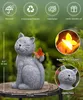 A gray cat figurine with an orange butterfly on its tail, placed on a wooden surface outdoors, with green foliage in the background. Insets show a close-up of the butterfly with LED light and a close-up of the cat's face. Produktbild-Vorschau