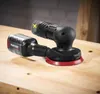 A black and red cordless orbital sander with a yellow 'X' marking on a wooden workbench. Produktbild-Vorschau