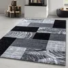 A modern abstract design rug with geometric patterns in black, gray, and white, placed on a light gray floor in a room with a fireplace and decorative logs. Náhled obrázku produktu