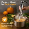 A hand is using a silver immersion blender to mix a soup in a pot, with pumpkins, lemons, and herbs in the background. Produktbild-Vorschau