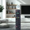 A remote control is held in the foreground, with a living room setting in the background, including shelves of books, a television, and furniture. Voir les images du produit
