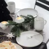 A festive table setting with Christmas decorations, including a black paper tree, pine branches, candles, and white ceramic tableware. Náhled obrázku produktu