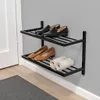 A two-tiered black metal shoe rack mounted on a gray wall, holding several pairs of shoes and two rolled-up towels. Produktbild-Vorschau