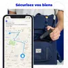 A person is holding a black GPS tracker attached to a keychain, with a blue backpack in the background. A smartphone screen shows a map with a route and alert. Náhled obrázku produktu