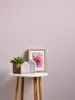 A small white table with a potted succulent, two decorative house-shaped objects, and a framed picture of a pink flower against a light pink wall. Produktbild-Vorschau