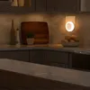 A kitchen counter with a cutting board, a bottle, a plant, and a bowl of fruit, with a night light plugged into the wall. Podgląd zdjęcia produktu