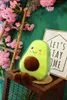 A plush avocado toy sits on a wooden surface, with pink flowers and a decorative sign in the background. Produktbild-Vorschau