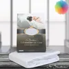 A product shot of a folded white fitted sheet with its packaging box behind it, set against a blurred background of a room. Produktbild-Vorschau