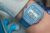 A close-up shot of a person's wrist wearing a blue Casio digital watch. A pair of blue and white earbuds are visible in the lower left corner. Náhled obrázku produktu