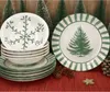 A set of Christmas-themed dinnerware with pine trees and snowflakes, arranged with decorative pinecones and a star. Produktbild-Vorschau