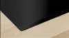 A close-up view of the edge of a black induction hob resting on a light wood countertop. Náhled obrázku produktu
