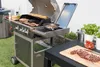 A barbecue grill is set up in an outdoor setting, with food being cooked and prepared on it and on a nearby table. Podgląd zdjęcia produktu
