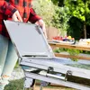 A person is opening a portable barbecue grill outdoors, with food on a table in the background. Produktbild-Vorschau