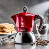 A red Bialetti Moka pot sits on a counter with toast and coffee beans nearby. Náhled obrázku produktu