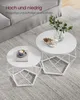 Two white round coffee tables with geometric bases are placed on a textured gray rug in a living room setting. A sofa is visible in the background. Produktbild-Vorschau