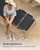 A woman is sitting on a black sun lounger on a sandy/gravelly surface. Náhled obrázku produktu
