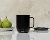 A black Ember mug sits on a white surface next to a charging coaster and two pears on a plate. Náhled obrázku produktu
