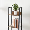 A close-up view of a tall, narrow bookshelf with wooden shelves and a black metal frame, displaying a potted plant on the top shelf and books and a decorative sculpture on the second shelf. Podgląd zdjęcia produktu