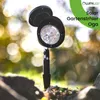 A black solar garden spotlight with a faceted lens is planted in the ground, with a blurred background of green foliage and bokeh lights. Produktbild-Vorschau