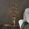 A decorative twig light set with LED lights in a glass vase, placed on a table next to a white armchair with a green throw. Podgląd zdjęcia produktu