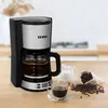 A coffee maker with a carafe full of coffee, a cup of coffee, a creamer pitcher, and coffee beans on a wooden table. Produktbild-Vorschau