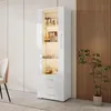 A white high-gloss display cabinet with glass doors and drawers, illuminated from within, stands in a modern room with a slatted wood accent wall and a plant. Produktbild-Vorschau