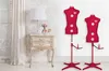 Two red dress forms on stands next to a vintage nightstand with a lamp and flowers. Produktbild-Vorschau