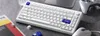 A white mechanical keyboard is shown on a gray surface, with a mouse, a video game controller, and a magazine nearby. Voir les images du produit