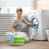 A woman is doing laundry in a laundry room, putting clothes into a washing machine. A green and white laundry basket is in the foreground. Produktbild-Vorschau