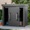 A dark gray metal storage shed with sliding doors, placed outdoors on a paved patio next to a building and surrounded by greenery. A blue watering can and a garden hose are visible inside the shed. Anteprima dell’immagine prodotto