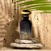 A perfume bottle is displayed on stones against a textured wall, with a palm leaf overhead. Podgląd zdjęcia produktu