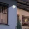 An outdoor LED floodlight with a motion sensor is mounted on the exterior wall of a building, illuminating the area. Produktbild-Vorschau