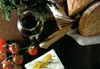 A rustic still life featuring bread, tomatoes, olive oil, and feta cheese, suggesting a meal preparation or serving scene. Produktbild-Vorschau