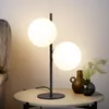 A modern black table lamp with two white glass globes sits on a round side table next to a bed. Náhľad obrázku produktu
