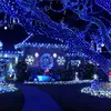 A house decorated with numerous blue and white Christmas lights, including lights on trees, the house itself, and a pathway leading to the house. A blue convertible with a figure in the driver's seat is parked in the yard. Náhled obrázku produktu