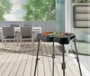 An electric grill with food on it is placed on a wooden deck next to a dining table and chairs, with a glass sliding door in the background. Náhled obrázku produktu