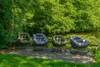 Four outdoor chairs are placed on a wooden deck next to a body of water, surrounded by lush green trees. Produktbild-Vorschau