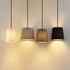 Four pendant lights with fabric shades in different colors hang against a light gray wall. Produktbild-Vorschau