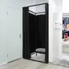 A black freestanding changing room with a black and white patterned stool inside, set up in a retail store environment next to a white door. Produktbild-Vorschau